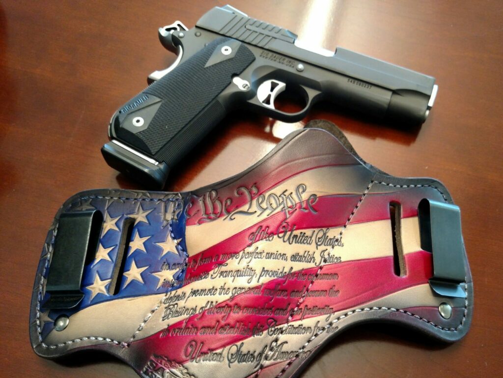 A 1911 pistol sits on a table above a leather holster